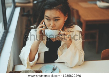 Young asian girl drinking coffee at cozy coffee shop and talking witch her friend via modern smartphone.