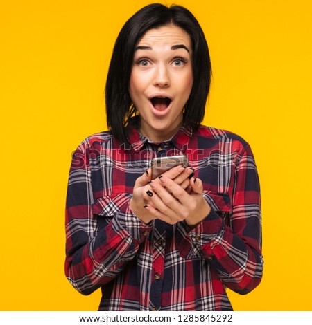 Excited laughing blogger woman in plaid shirt standing and using mobile phone over yellow background - Image