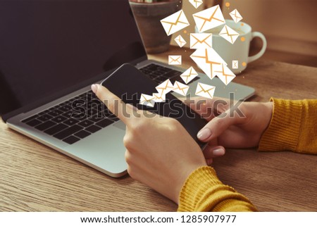 cropped image of woman holding smartphone with email icons at table with laptop