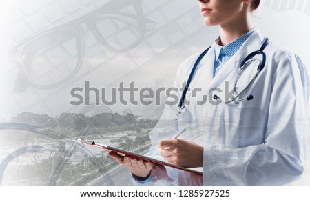 Portrait of young female doctor in white sterile suit holding notebook in hands while standing against modern city view and medical equipment on background. Double exposure