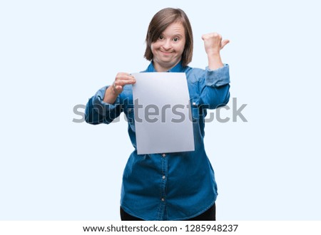Young adult woman with down syndrome holding blank paper sheet over isolated background pointing and showing with thumb up to the side with happy face smiling