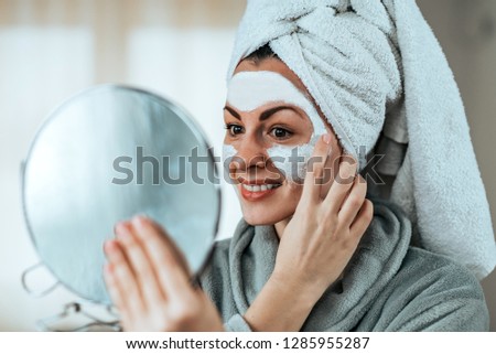 Smiling woman holding a mirror in hand and applying cosmetic facial mask, close-up.