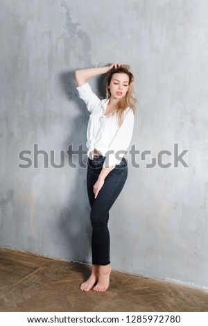 Model blonde blue jeans white shirt standing gray wall by window  copy space. Openness comfort portrait free woman thinks decides chooses. Posing emotions.