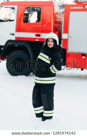 Photo of young firefighter woman staring into camera with black hair next to fire engine. Smirked happy portrait of a woman firefighter