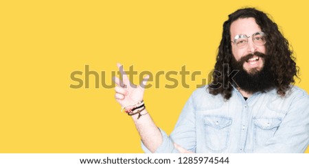 Young hipster man with long hair and beard wearing glasses with a big smile on face, pointing with hand and finger to the side looking at the camera.