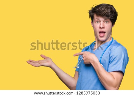 Young doctor wearing medical uniform over isolated background amazed and smiling to the camera while presenting with hand and pointing with finger.