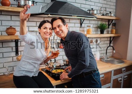 Cheerful couple stand together in kitchen. She sit at stove and take selfie. Guy pose and fry food. He smile.