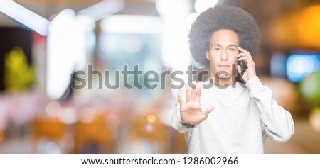Young african american man with afro hair talking on smartphone with open hand doing stop sign with serious and confident expression, defense gesture
