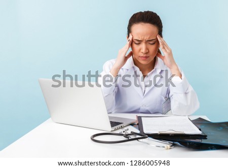Photo of a young tired bored woman doctor with headache posing isolated over blue wall background using laptop computer.