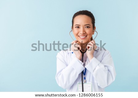 Photo of a young cheerful happy woman doctor posing isolated over blue wall background with stethoscope.