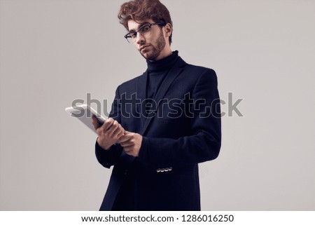Fashion portrait of handsome elegant man with curly hair wearing suit and glasses holding a tablet on gray background in studio