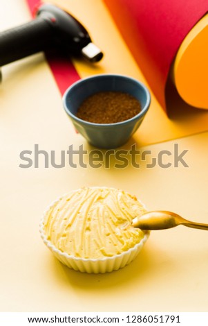 Rows of Creme catalana or creme brulee prepared to be caramelized, with a golden white spoon, a blowtorch and brown cane sugar on a yellow tableboard.