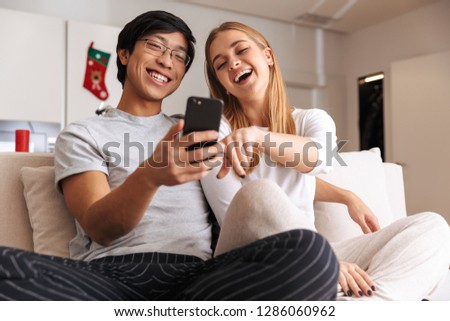 Cheerful young couple sitting together on a couch at home, looking at mobile phone