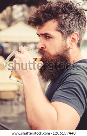 Man with beard and mustache holds glass with beer while relaxing at cafe terrace. Guy having rest with cold draught beer. Hipster on calm face drinking beer outdoor. Draught beer concept.