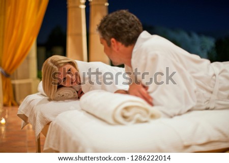 Smiling husband and wife lying on massage tables set up on a candlelit patio.
