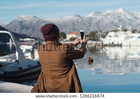 Woman in a beige coat makes photo by the sea and Snow-capped mountains on the background of Montenegro