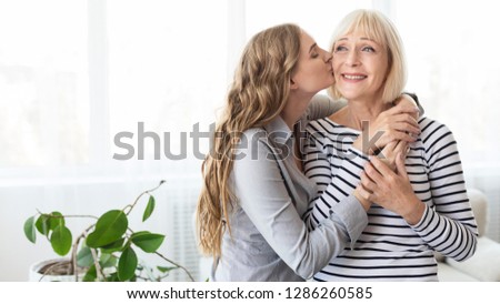 Daughter kissing her mature mother on the cheek, standing against window at home