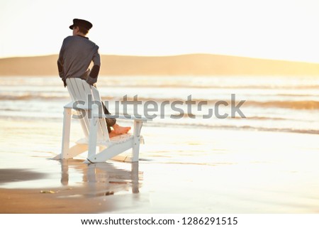 Boy looks along shoreline as he stands on the seat of an Adirondack chair sitting on a beach.