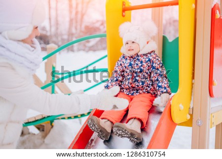 Winter children games on street. Little girl riding down hill Playground outdoors, mom gives hand