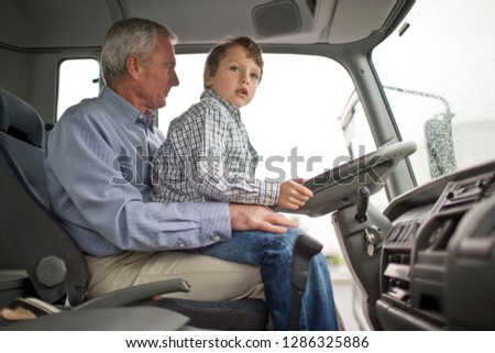 Young boy sitting on his father's lap behind the steering wheel of a truck.