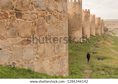 Asian girl wearing sunglasses, a scarf and wellington boots, looks away as she walks beside the Walls of the old town of Avila, a national monument and world heritage site in Castilla y Leon, Spain