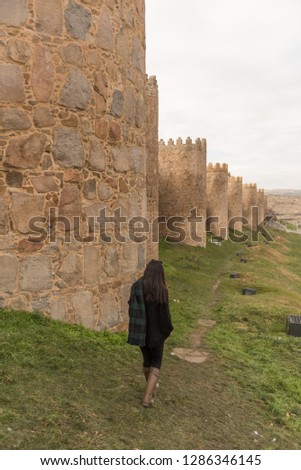 Asian girl wearing sunglasses, a tartan scarf and wellington boots, walks beside the Walls of the old town of Avila, a national monument and world heritage site in Castilla y Leon, Spain