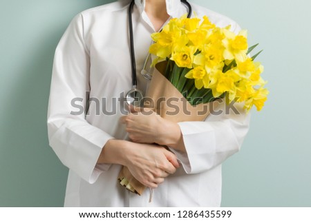 Closeup of bouquet yellow flowers in hands of female doctor, pastel green mint wall background. World health day, doctors day. Royalty-Free Stock Photo #1286435599