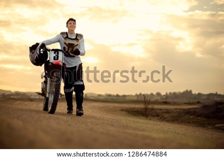 Smiling young female motocross rider pushing her dirt bike at the track.