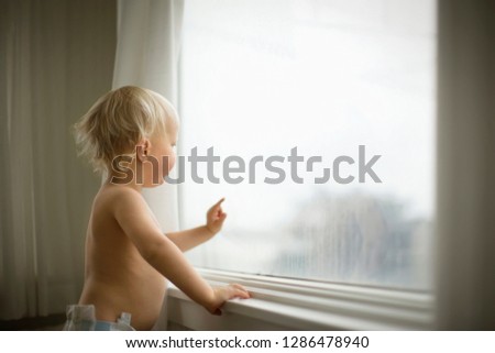 Shirtless young toddler looking out the window of his home.