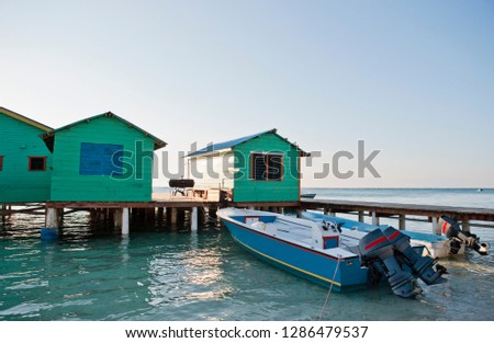 Moored boats next to boatsheds on a jetty in the sunshine.
