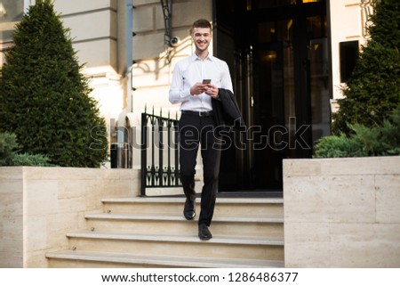 Young smiling businessman in white shirt with wireless earphones holding black jacket and cellphone in hand happily looking in camera going down the stairs with beautiful building on background