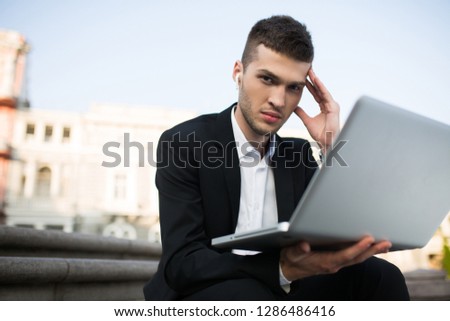 Young serious businessman in classic black jacket and white shirt with wireless earphones thoughtfully looking in camera with laptop in hand outdoor