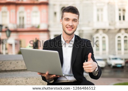 Young smiling businessman in classic black suit and white shirt with wireless earphones joyfully looking in camera showing thumb up gesture with laptop in hand outdoor
