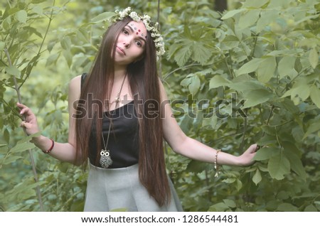 A forest picture of a beautiful young brunette of European appearance with dark brown eyes and large lips. On the girl's head is wearing a floral wreath, on her forehead shiny decorations