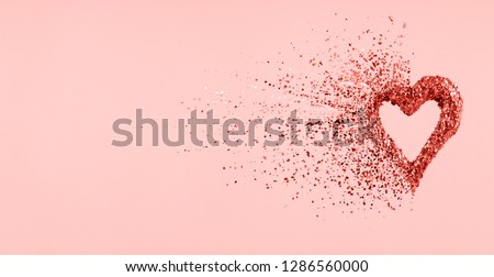 Glitter heart dissolving into pieces on pink background.  Valentines day, broken heart and love emergence concept. Horizontal wide screen banner format, living coral toned - color of the year