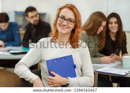 Portrait of young red haired girl with blue folder in hands. Happy beautiful student in glasses smiling in front of camera. Pupils sitting on lecture from foreign language in private school.