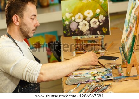 Creativity man with bread wearing uniform, working alone at workplace with pains and brushes. Male artist with perfect imagination drawing picture looking down on palette. Professional painter.