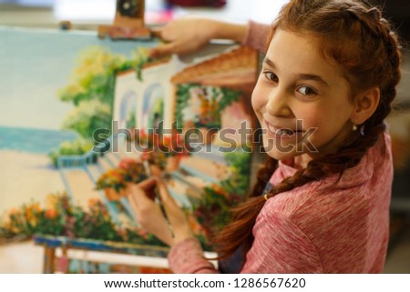 Happy little red haired girl with braid drawing beautiful picture of sea view and showing it. Pretty female pupil studying of classic art, smiling at camera. Concept of art and child hobby.