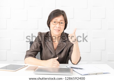 Portrait of Asian, senior businesswoman, short hair, wearing eyeglasses, sitting in meeting room with notebook, smartphone, notepad, tablet , making thumb up sign with confidence