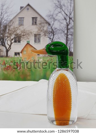 Handmade decorative figure of melted glass duck with a photo background of house and paper cutting craft in form of little house. Rural life concept.