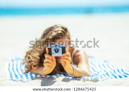 Closeup on retro photo camera holding by woman laying on a striped towel on the seashore. Capture vacation in incredible photos preserve those memories and share with family and friends & on social.