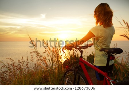 Young lady standing with bicycle on a sea coast and enjoying sunset Royalty-Free Stock Photo #128673806