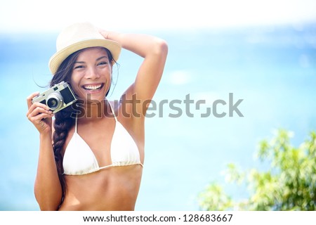 Beach fun people - woman with retro vintage camera playful laughing in bikini on blue ocean background wearing beach hat. Multicultural Asian / Caucasian girl.