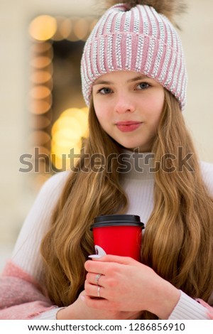 A girl in a pink warm jacket and hat with 
pompon stands with coffee in the hand on the background of golden yellow lights and blurred shop windows. Winter wonderland. Christmas and New Year concept.