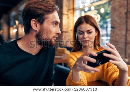 Red-haired woman shows something to a man in a phone sitting at a table in a cafe                      
