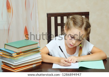 Little girl sitting at table with pile of books and doing homework