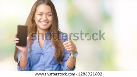 Young beautiful brunette business woman showing screen of smartphone over isolated background happy with big smile doing ok sign, thumb up with fingers, excellent sign