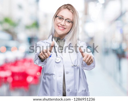 Young blonde doctor woman over isolated background approving doing positive gesture with hand, thumbs up smiling and happy for success. Looking at the camera, winner gesture.