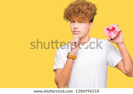 Young handsome man holding heart box as gift of valentine's day serious face thinking about question, very confused idea