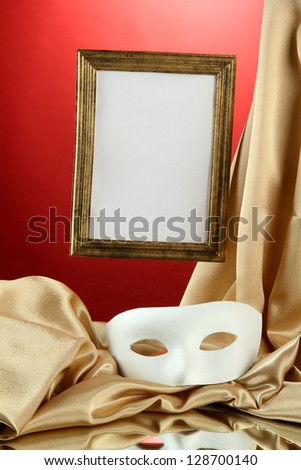 White mask, empty frame and golden silk fabric, on red background